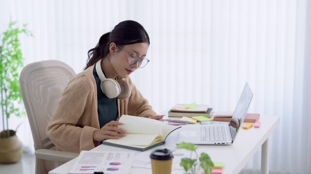Young Asian woman working remotely on laptop at home office desk with headphones and documents