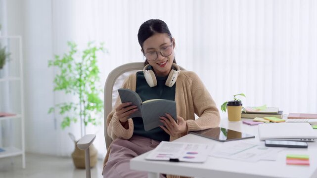 Young Asian Gen Z professional reading notebook with smartphone while studying at bright modern home workspace