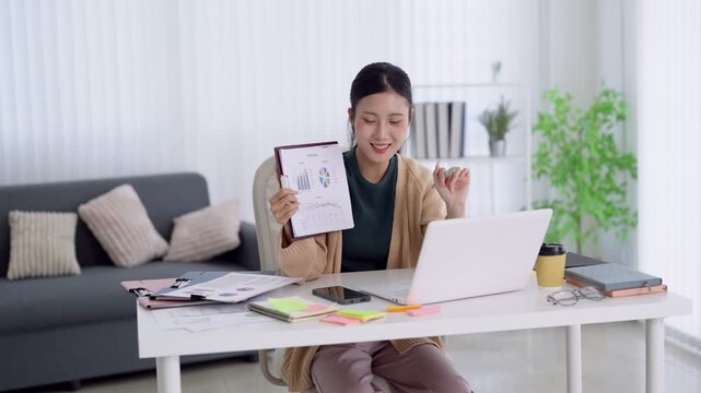 Young Asian professional presenting business analytics report during video conference from bright home office workspace