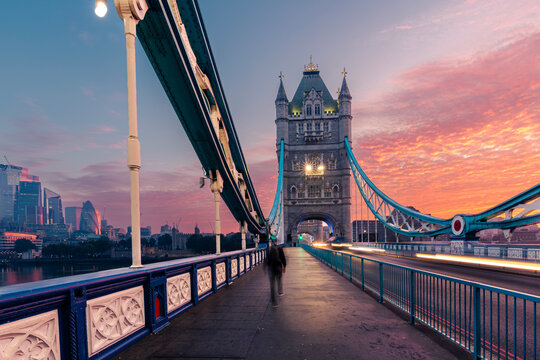 Motion blur silhouette of mid adult passenger walking across Tower Bridge in London during dramatic morning sunrise. European travel destination, historic city commute and transportation concept.