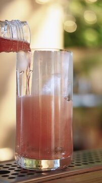 Vertical video. Cocktail making. Pouring drink. Adding refreshing flavored pink soda liquid in glass with ice cube from bottle on pub counter.