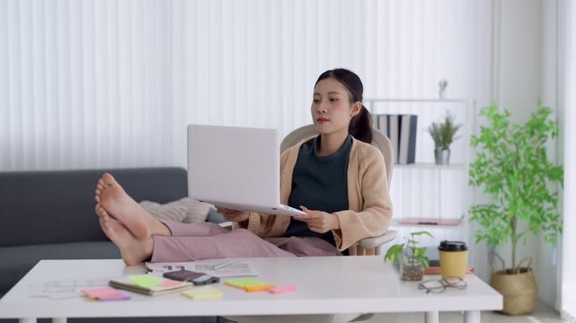 Young Asian woman working remotely from home with laptop feet up on table in bright modern living room