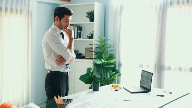 Professional civil engineer standing and drinking coffee and thinking about house plan at meeting table with equipment scatter around. Stressful project manager looking at blueprint. Alimentation.