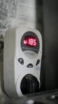 Close-up vertical view of a white voltage protector plugged into a socket with a red LED display showing numbers. Concept of home electrical safety, power surge protection, and unstable energy