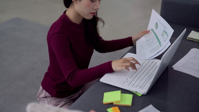 Young Asian Gen Z professional analyzing financial report while working remotely on laptop at modern home office desk