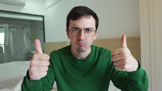Serious man with glasses showing two thumbs up with a disapproving facial expression, conveying sarcasm and passive aggression while sitting in a bright room with a bed visible