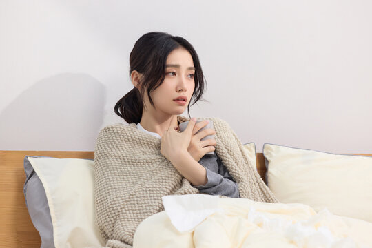 Young woman feeling unwell in bed with blanket and hot drink at home