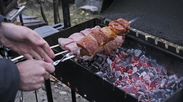 A man arranges raw marinated pork skewers over red-hot charcoal embers outdoors. The close-up shot captures the grilling process and the smoky barbecue atmosphere in the garden.