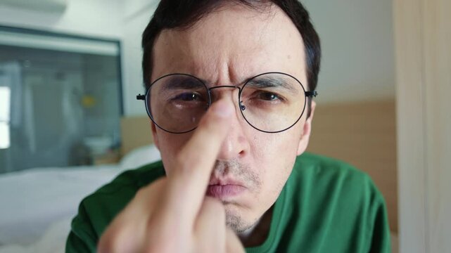Close-up portrait of an angry man flipping off the camera by adjusting his glasses with the middle finger. Frowning guy showing offensive hidden gesture of contempt and disrespect