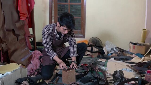 A focused man crouches at a cluttered indoor workbench, examining tools and assembling parts amid wires, wood planks, and scattered materials in a dimly lit