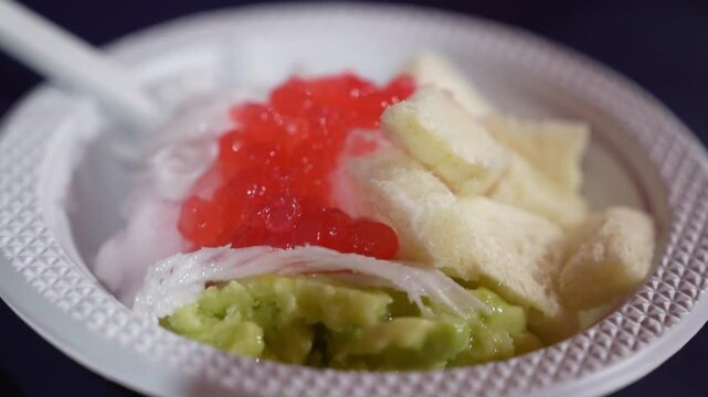 Close-up of Indonesian es campur dessert with shaved ice, sweet syrup, avocado, jelly, and crispy toppings in a bowl, refreshing street food dessert footage.