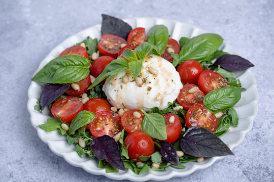 Fresh burrata salad served with ripe cherry tomatoes, mixed greens, fresh basil leaves, and toasted pine nuts on a gray background. Healthy eating concepts, and Italian cuisine. side view