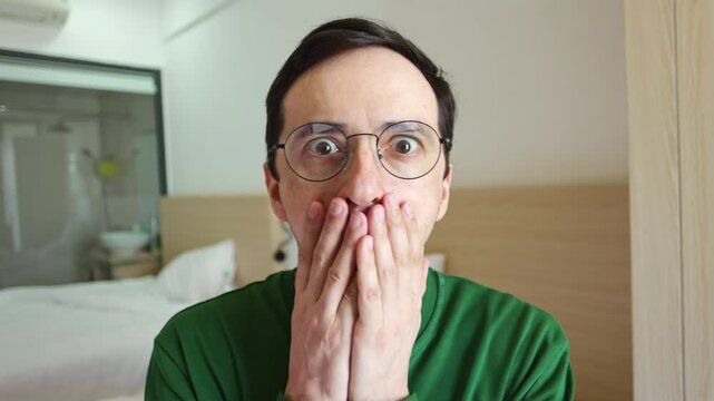 Young man wearing eyeglasses looking directly at the camera with a shocked expression, his eyes wide and mouth open in disbelief as he covers it with both hands in a bedroom interior setting