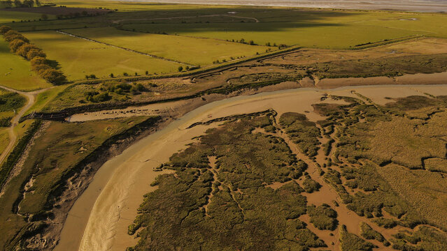 Aerial view of a winding tidal creek and salt marshes bordered by green agricultural fields in the Borough of Swale, England, United Kingdom.