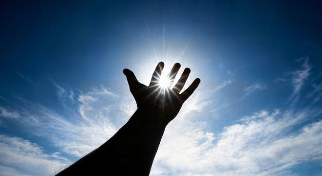 Silhouette of outstretched hand reaching towards bright shining sun in blue sky with white clouds, symbolizing hope and aspiration.