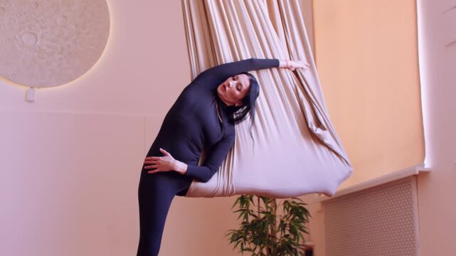 Body exercise. Fitness studio. Female doing side stretch in split pose with leg support on fly yoga hammock for spine strength.