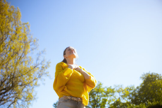 A calm woman rests her hands on her heart in a serene park setting, practicing mindfulness and self-care to cultivate inner peace and stress relief.