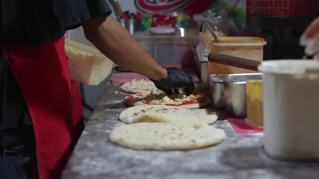 Chef preparing flatbread pizza with tomato sauce and shredded cheese in a commercial kitchen, close-up restaurant food preparation footage.