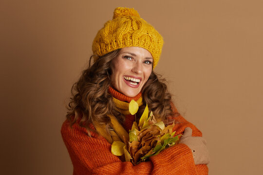 A smiling woman in her 30s, adorned in a yellow knit hat, orange sweater, and scarf, holds a vibrant bouquet of autumn leaves against a brown background.