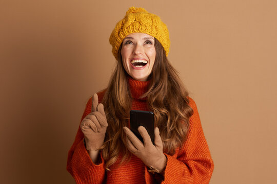 Cheerful woman in yellow beanie and orange sweater smiles broadly, looking upwards with index finger pointed, holding smartphone. She embodies eureka moment of inspiration or exciting new idea