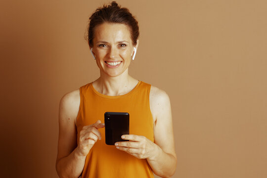 A smiling adult woman with hair in a bun and wireless earbuds points at her smartphone screen. She wears an orange-yellow top and looks at the camera, conveying cheerful digital engagement.