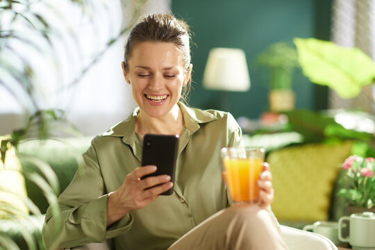 Looking down at her smartphone with a smile, a woman relaxes on her sofa with a glass of orange juice, surrounded by plants and sunlight.