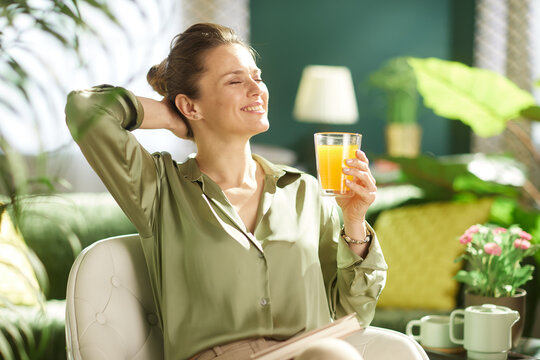 A woman sits comfortably in her sunlit living room, smiling with her eyes closed while holding a glass of juice, embracing a moment of calm.