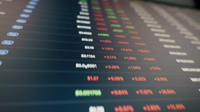 Digital screen displays scrolling cryptocurrency and stock market data with changing prices. The shallow depth of field and angled perspective create a dynamic, modern financial technology feel.