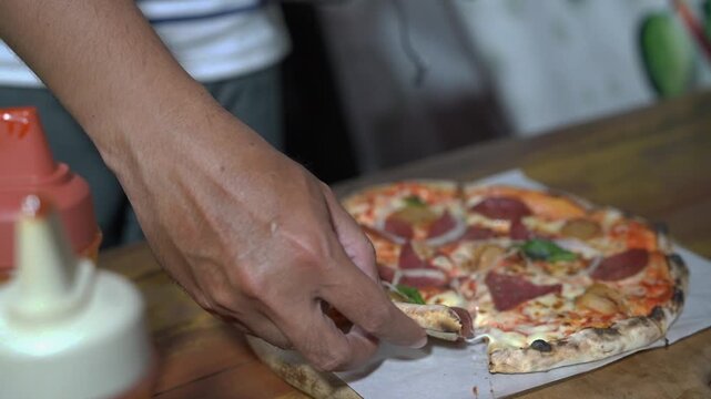 Close-up of a hand lifting a cheesy pepperoni pizza slice from a wooden board, showing melted cheese pull in a casual restaurant setting.