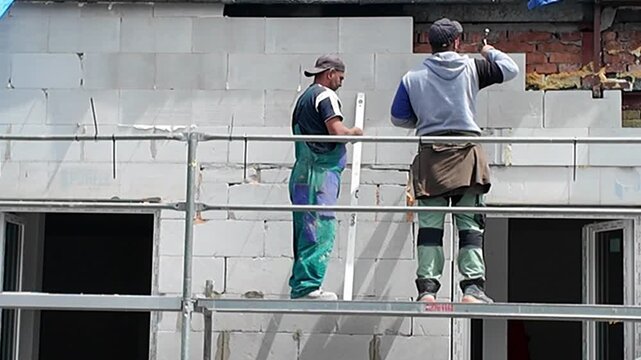 Construction workers building a wall with cinder blocks on scaffolding. Workers in protective gear meticulously construct a wall using large cinder blocks, demonstrating precision and teamwork on a