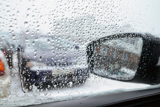 A close-up view through a car window covered in raindrops and melting snow, framing a black side-view mirror that reflects a red brick building on a wet, overcast winter day.