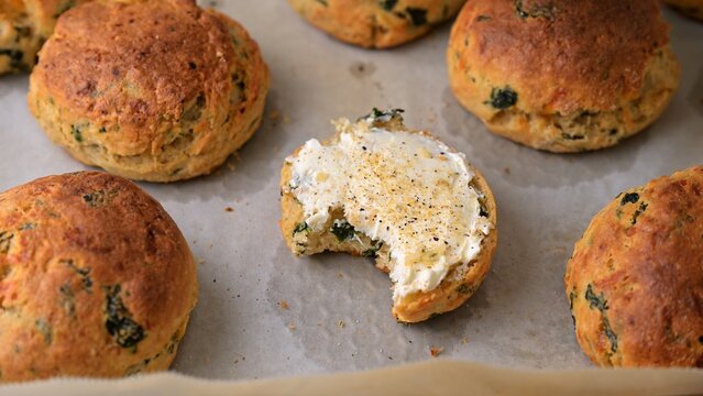 Homemade spinach and parmesan savory biscuits on parchment paper, one cut open and topped with cream cheese and spices. Freshly baked cheesy herb biscuits, close-up food photography.