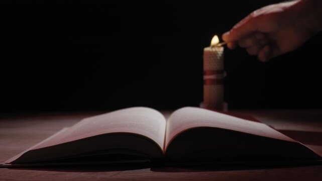 A hand with a match lights a beeswax candle, casting a warm, flickering glow over an open book on a wooden table. Concept of seeking knowledge, tradition, or finding hope in darkness.