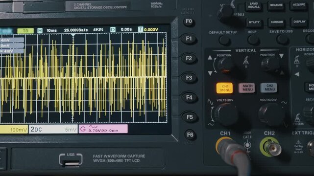 A digital oscilloscope screen displays a complex yellow electrical waveform. The camera slowly zooms out to reveal the control panel and measuring equipment. Concept of data analysis and engineering.