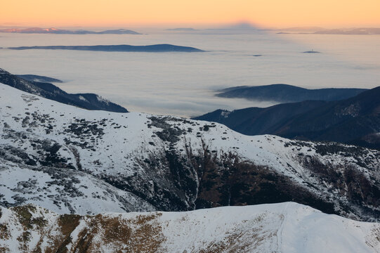 Aerial view of Western Tatras mountains covered in snow with a sea of clouds below during golden hour in Tatra County, Malopolskie, Poland.