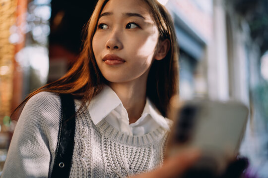 Close-up portrait of Asian woman looking aside, smartphone in hand. Expression conveys inner thoughts, situational awareness, and digital presence in an urban setting.