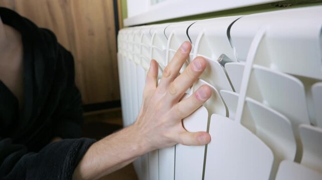 A man in a black robe places his hands on a white central heating radiator to check the temperature indoors. Concept of winter season, cold weather, energy crisis, and utility bills.