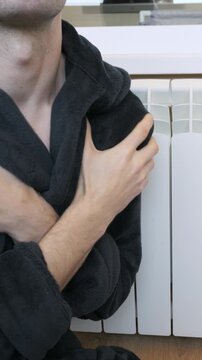 A man in a warm bathrobe hugs himself and shivers while sitting next to a white radiator. Concept of heating problems, cold apartment, and high energy bills during winter season.