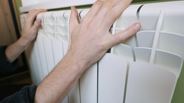 A man's hands touch a white heating radiator, checking the temperature and warmth in the room. Concept of the heating season, home comfort, rising energy prices, and saving heat.