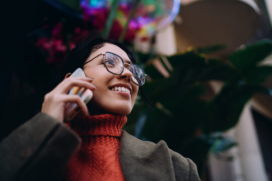 Close up of happy mixed race woman talking on cellphone, wearing glasses and orange turtleneck, urban setting, joyful emotion, wireless communication, mobile app user, authentic lifestyle.