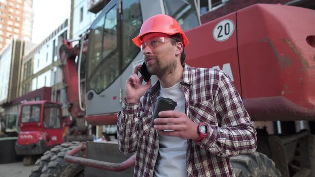 Smiling builder in a hard hat enjoys a coffee break and talks on the phone at a construction site. Happy foreman near heavy machinery communicating with team. Satisfied professional at work