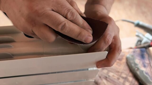 Hands use a sandpaper tool on a plastic item in a workshop. The person is preparing the surface for a project. Focus is on the work being done and the tools in use.