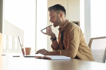 Fototapeta premium Focused Caucasian Male Working From Home, Using Laptop And Voice Recorder While Sitting At Desk In Modern Office. Professional Atmosphere With A Concept Of Remote Working And Productivity.
