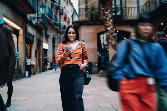 Smiling Hispanic woman walking on city street using smartphone. Young dark skinned model in orange cardigan browsing social media. Authentic representation of digital lifestyle and urban fashion.