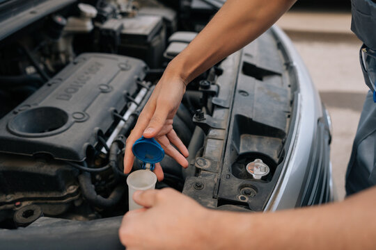 Close-up shot of auto mechanic performing vehicle maintenance. Unrecognizable person opens blue cap of washer fluid reservoir to check liquid level in engine compartment. Focus on hands and action.