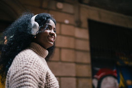 Cheerful African American woman in headphones profile portrait, dark skinned female looking away with smile, urban music listener, wireless audio gadget, 5G internet connection, city lifestyle.