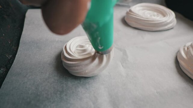 Pastry chef squeezes thick white meringue cream from a green piping bag onto parchment paper. Sweet dessert preparation process takes place in a home kitchen.