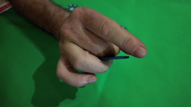 A close-up shot of an adult man's hands handling and manipulating a blue poker chip with the number 25 on a green felt table. Other poker chips of various colors are visible in the background