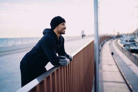 Smiling athlete resting on bridge railing at marina, man in winter sportswear enjoying city harbor view, positive attitude and mental health, urban wellness routine, healthy life in city.