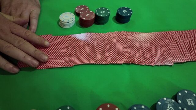 A close-up view of hands spreading out a full deck of poker cards face-up on a vibrant green table, revealing various card values and suits. Several stacks of colorful poker chips are visible in the